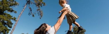 happy mother lifting toddler daughter against green trees and clear sky, banner  clipart
