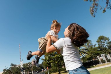 Mutlu anne, küçük kızını yeşil ağaçlara ve bulutsuz gökyüzüne karşı kaldırıyor. 
