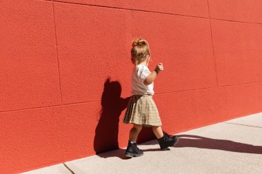Kareli etekli ve beyaz tişörtlü küçük kız kırmızı duvarlı binanın yanında yürüyor. 