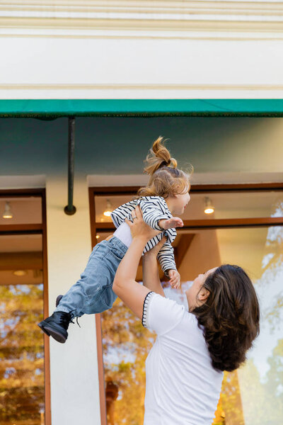 Брюнетка мать поднимает дочь в полосатой рубашке с длинным рукавом возле витрины магазина в Майами 