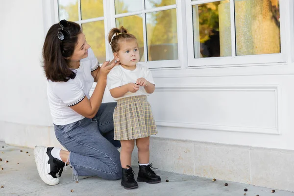 stock image happy mother in jeans looking at toddler daughter in skirt near house 