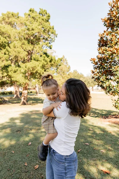 brunette mother kissing baby daughter while standing together in park of Miami