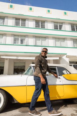MIAMI, FLORIDA, ABD - 15 Aralık 2022: klasik retro arabanın kapısını açan güneş gözlüklü mutlu Afrikalı Amerikalı adam 