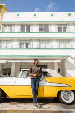 MIAMI, FLORIDA, ABD - 15 Aralık 2022: Klasik sarı arabanın yanında duran mutlu Afro-Amerikan adam 