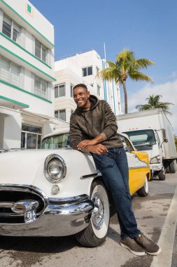 MIAMI, FLORIDA, ABD - 15 ARALIK 2022: Eski model arabanın yanında duran mutlu Afrikalı Amerikalı adam 