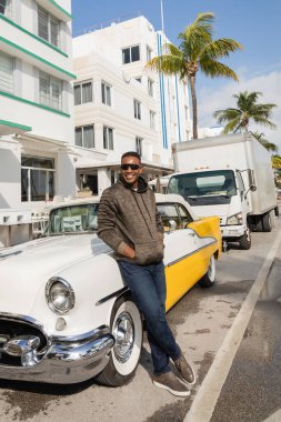 MIAMI, FLORIDA, ABD - 15 Aralık 2022: Güneş gözlüklü neşeli Afrikalı Amerikalı adam klasik arabanın yanında duruyor 