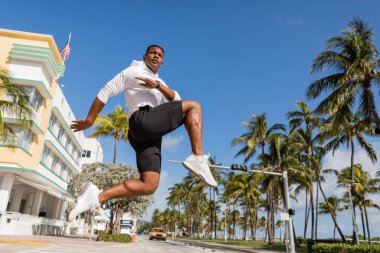 sportive african american man jumping next to palm trees and modern condominium in Miami  clipart
