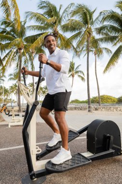 Neşeli Afrikalı Amerikalı adam dışarıda spor salonunda çapraz antrenörle egzersiz yapıyor. 
