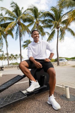 Kaygısız Afro-Amerikan sporcusu Miami 'deki spor salonunda çapraz antrenör ekipmanının üzerinde oturuyor.