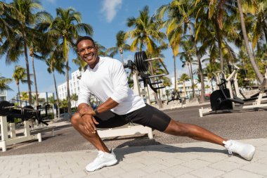 full length of happy african american sportsman stretching in outdoor gym in Miami  clipart