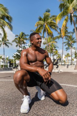 happy african american sportsman in shorts and sneakers smiling after workout in Miami beach  clipart