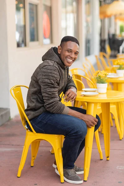 happy african american man in hoodie and jeans sitting at bistro table in outdoor cafe in Miami