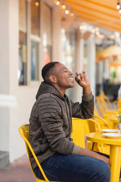 stock image side view of pleased african american man in hoodie and jeans sitting at bistro table in outdoor cafe 