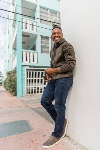 полная длина веселый африканский американец в толстовке проведение стильные солнцезащитные очки, стоя на открытом воздухе 