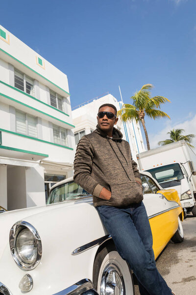 MIAMI, FLORIDA, USA - DECEMBER 15, 2022: african american man in sunglasses standing with hands in pockets near vintage car 