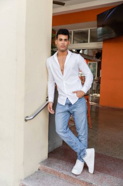 Full length of cuban man in white shirt and jeans posing in outdoor cafe on street in Miami clipart