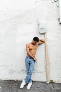 Shirtless cuban man in jeans standing near wire on white building in Miami during summer, muscular  clipart