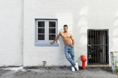Kot pantolon ve gözlüklü üstsüz genç bir adam Miami 'de beyaz duvarın yanında duruyor.