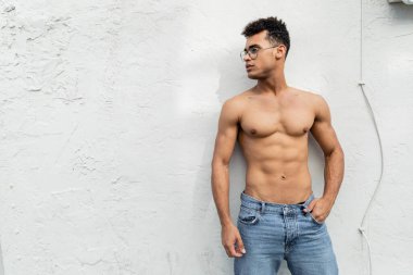 cuban man with athletic body posing in blue jeans and trendy round-shaped eyeglasses near building clipart