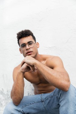 Portrait of relaxed and muscular young cuban man in trendy round-shaped eyeglasses and blue jeans  clipart