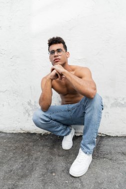 Shirtless and young cuban man with athletic body wearing round-shaped eyeglasses and blue jeans clipart