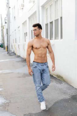 Full length of confident and muscular young cuban man in round-shaped eyeglasses and jeans  clipart
