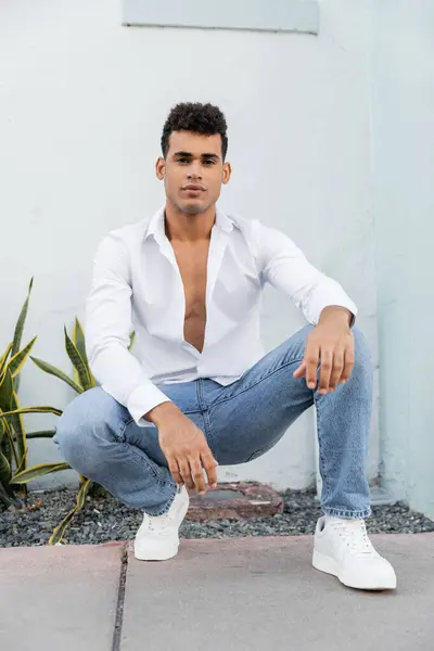 stock image cuban man in trendy white shirt and jeans posing and looking at camera near building in Miami