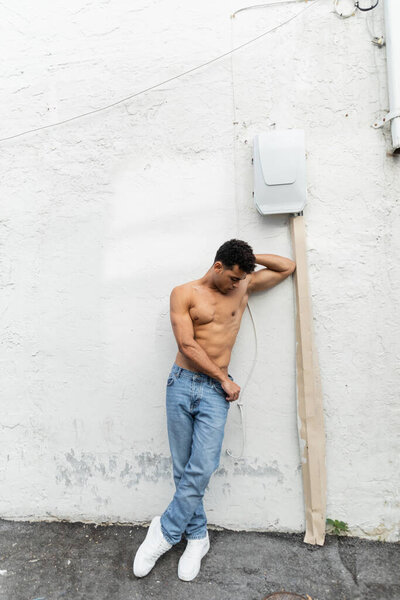 Shirtless cuban man in jeans standing near wire on white building in Miami during summer, muscular 
