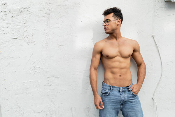 cuban man with athletic body posing in blue jeans and trendy round-shaped eyeglasses near building