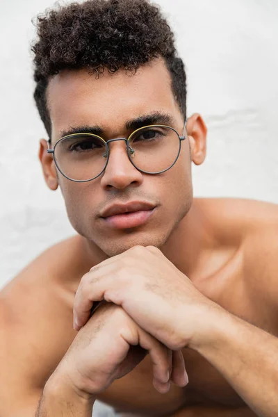 stock image Portrait of curly and shirtless cuban man in stylish round-shaped eyeglasses holding hands near chin