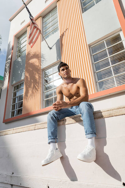 Low angle of cuban man with muscular body posing in baseball cap and jeans in Miami, south beach