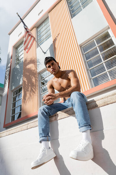 cuban man with muscular body posing in baseball cap and jeans on street in Miami in summer day