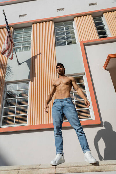 muscular cuban man in baseball cap and jeans standing on parapet near building with american flag 