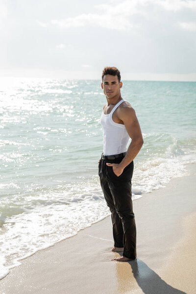 young cuban man in wet clothes holding hand in pocket of pants, standing on sand near ocean on Miami