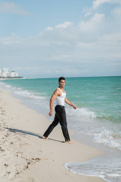 Muscular cuban young man walking on sand near ocean water of Miami South Beach, Florida