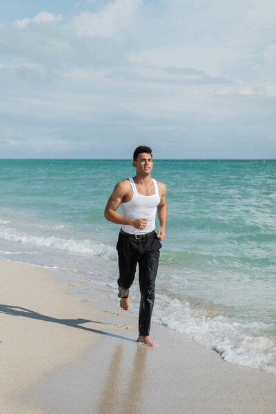 Muscular cuban young man running on sand near ocean water of Miami South Beach, Florida