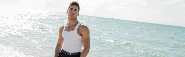 stylish young cuban man looking at camera with ocean water and sky at background in Miami, banner 