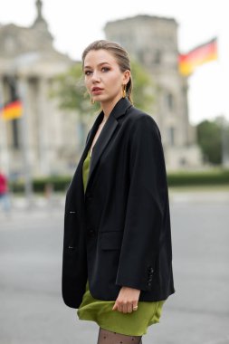 Portrait of young fair haired woman in fashionable silk dress and black jacket on street in Berlin clipart