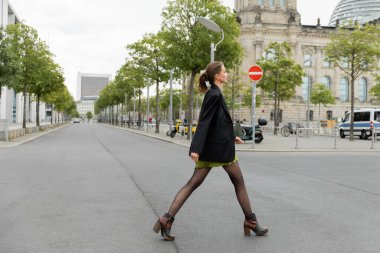 Side view of fashionable young woman in dress and black jacket walking on road in Berlin, Germany clipart