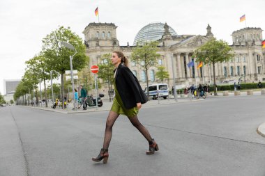 Full length of young and fair haired woman in silk dress, jacket and boots walking on road in Berlin clipart