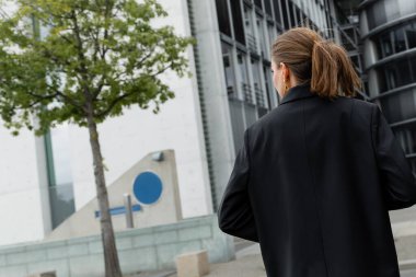 Berlin, Almanya 'da siyah ceketli, şık ve açık tenli genç bir kadının arka planda yürüyüşü 