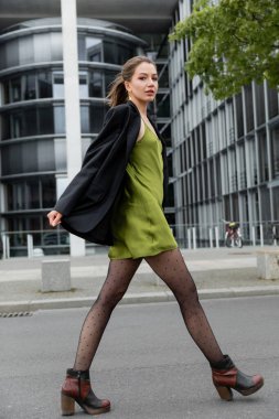 Yeşil ipek elbiseli, puantiyeli tayt ve ceketli şık bir kadın Berlin 'de kameraya bakıyor.