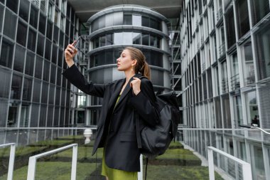 Sırt çantalı ve akıllı telefondan selfie çeken şık, genç bir kadının yan görüntüsü.  