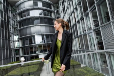 Side view of cheerful young fair haired woman in black jacket and silk dress standing in Berlin clipart