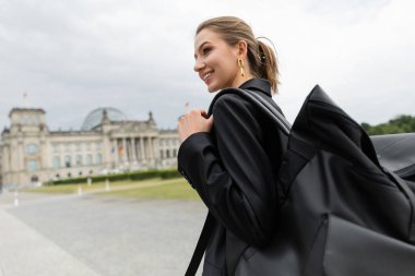 Cheerful woman in jacket and dress holding backpack while walking near Reichstag Building in Berlin clipart
