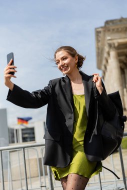 Cheerful woman in jacket and silk dress holding backpack and taking selfie  in Berlin, Germany  clipart