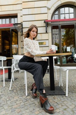 Dantelli bluz ve pantolonlu şık bir kadın kahve tutuyor ve Berlin 'deki açık hava kafeteryasında oturuyor. 