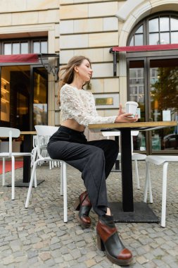 Dantelli bluzlu, pantolonlu, kahve tutan ve Berlin 'deki açık hava kafeteryasında oturan rüya gibi bir kadın. 