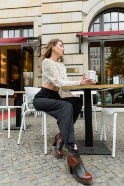 dreamy woman in lace top and pants holding coffee and sitting at table of outdoor cafe in Berlin  clipart