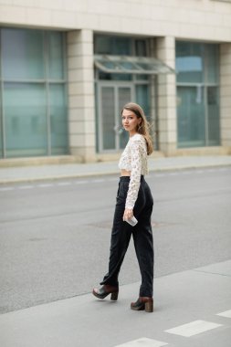 Dantelli bluz ve pantolonlu, elinde kahve ile kameraya bakan, uzun boylu, modaya uygun bir kadın.